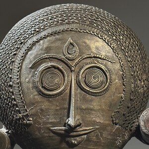 May include: A bronze sculpture of a face with intricate details. The face has large, round eyes with spiral patterns, a prominent nose, and a small mouth. The sculpture is decorated with a patterned design around the head.