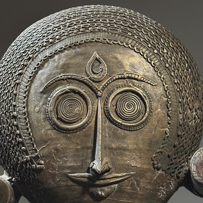May include: A bronze sculpture of a face with intricate details. The face has large, round eyes with spiral patterns, a prominent nose, and a small mouth. The sculpture is decorated with a patterned design around the head.