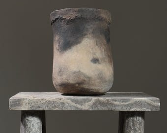 Puede incluir: Un jarrón de cerámica rústico, hecho a mano, con un esmalte moteado marrón y blanco. El jarrón está sentado sobre un pedestal de piedra con una superficie rugosa y texturizada.