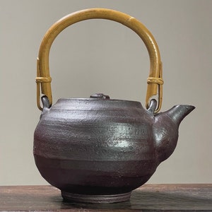 Può includere: Una teiera in ceramica marrone scuro con un manico in bambù. La teiera ha un corpo rotondo e un beccuccio laterale.