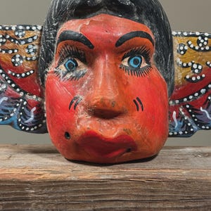 May include: A hand-painted wooden mask with a red face, black hair, and blue eyes. The mask has large, expressive features and is decorated with colorful wing-like elements on either side. The mask is resting on a wooden surface.