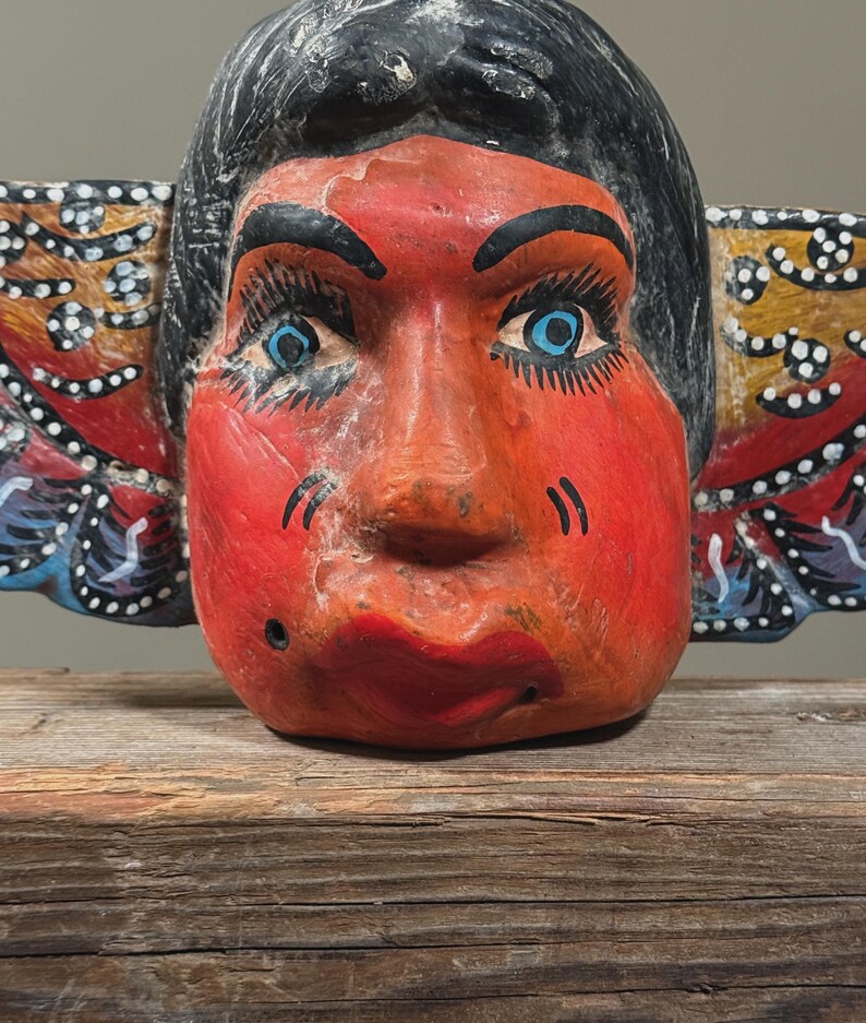 May include: A hand-painted wooden mask with a red face, black hair, and blue eyes. The mask has large, expressive features and is decorated with colorful wing-like elements on either side. The mask is resting on a wooden surface.