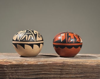 Puede incluir: Dos cuencos de cerámica pintados a mano con diseños geométricos en blanco y negro. El cuenco de la izquierda es beige con diseños negros, y el cuenco de la derecha es rojo con diseños negros. Los cuencos están sobre una superficie de madera.
