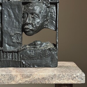 May include: A black abstract sculpture of a face and body, partially obscured by geometric shapes. The sculpture is made of a rough, textured material and is displayed on a light gray stone base.