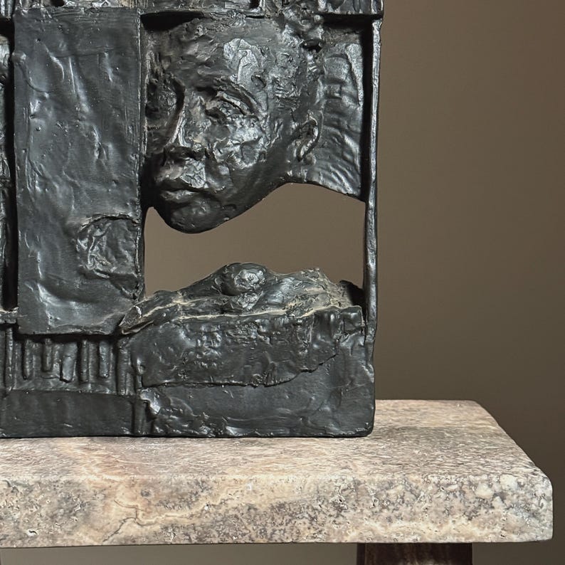 May include: A black abstract sculpture of a face and body, partially obscured by geometric shapes. The sculpture is made of a rough, textured material and is displayed on a light gray stone base.