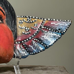 May include: Close-up of a colorful, hand-painted wooden bird sculpture. The bird features a red body, black head, and a detailed wing with yellow, red, and blue sections, accented with white dots. The sculpture is displayed on a clear stand.