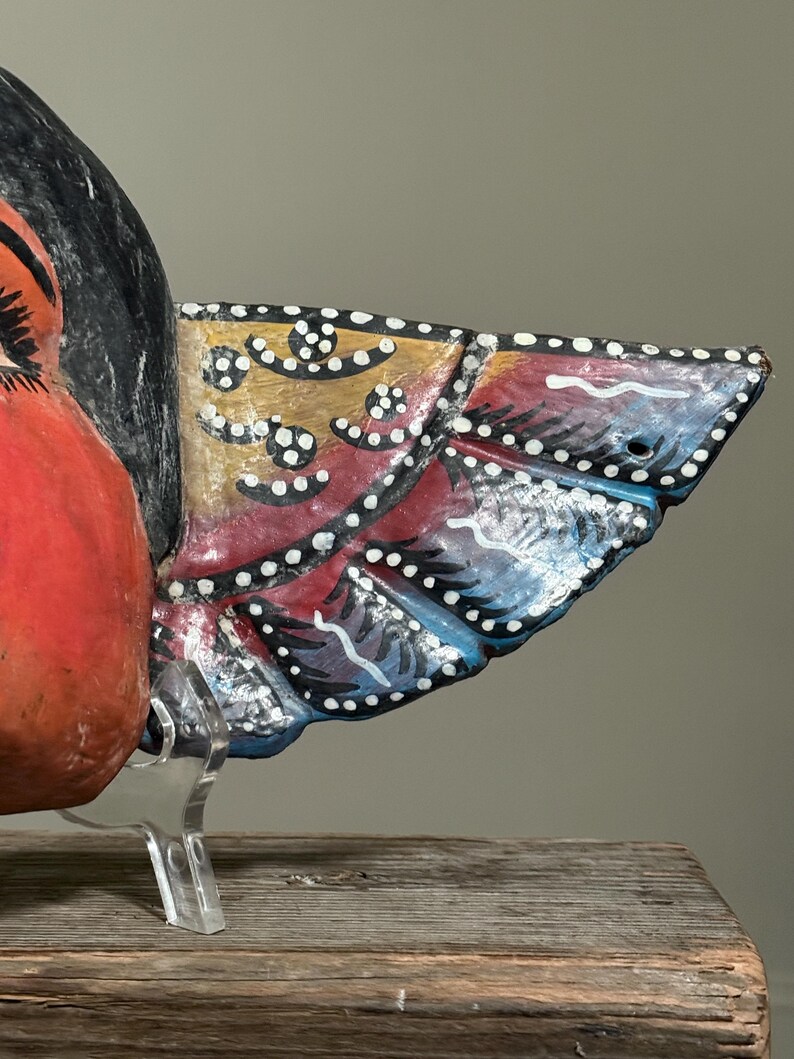 May include: Close-up of a colorful, hand-painted wooden bird sculpture. The bird features a red body, black head, and a detailed wing with yellow, red, and blue sections, accented with white dots. The sculpture is displayed on a clear stand.