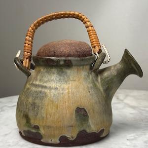 May include: A ceramic teapot with a brown, green, and yellow glaze. The teapot has a woven wicker handle and a cork lid.