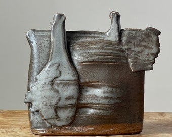 May include: A ceramic vase with a textured, gray and brown glaze. The vase has an abstract, organic shape with multiple protrusions.