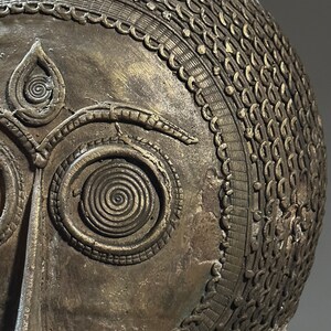 May include: A bronze mask with intricate details, including a spiral design around the eyes and a patterned border around the top of the head.