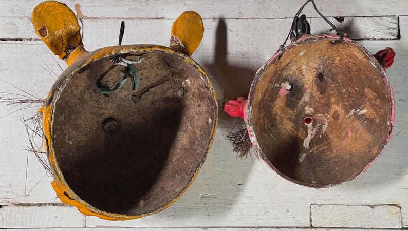 Puede incluir: Dos m&aacute;scaras vintage de papel mach&eacute; con detalles pintados. Una m&aacute;scara es amarilla con detalles marrones y verdes, la otra es roja con detalles marrones. Ambas m&aacute;scaras tienen un interior hueco.