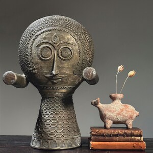 May include: A gold-toned, decorative sculpture with a human-like face and a patterned body. The sculpture is standing on a wooden surface. A small, terracotta sculpture of a stylized animal is in the foreground, sitting on a stack of books.
