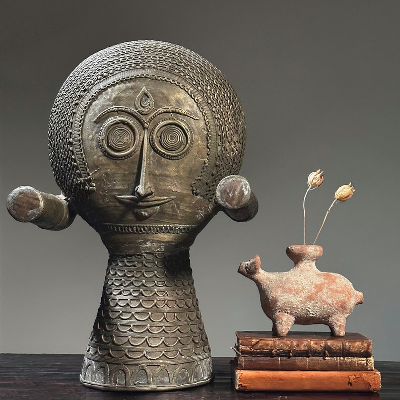 May include: A gold-toned, decorative sculpture with a human-like face and a patterned body. The sculpture is standing on a wooden surface. A small, terracotta sculpture of a stylized animal is in the foreground, sitting on a stack of books.