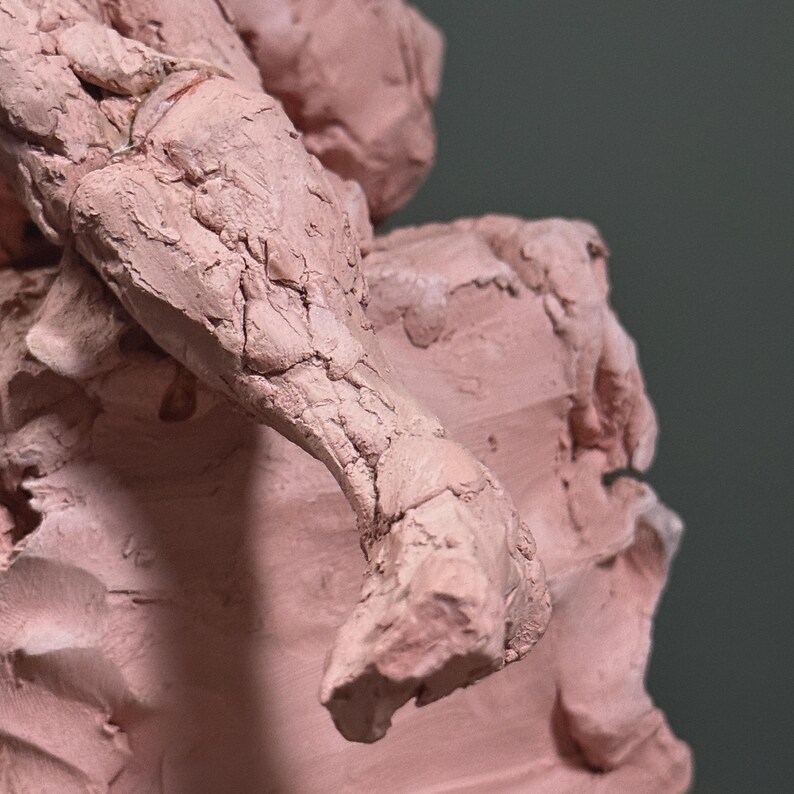 May include: Close-up of a clay sculpture in a reddish-brown hue. The arm and part of the torso are visible, with detailed textures and cracks. The sculpture is set against a muted green background, highlighting the form and texture of the artwork.