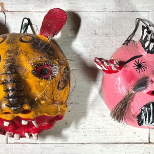 Puede incluir: Dos m&aacute;scaras de papel mach&eacute; hechas a mano. Una m&aacute;scara es amarilla con detalles negros y rojos y tiene un hocico largo. La otra m&aacute;scara es rosa con detalles negros y blancos y tiene un bigote.