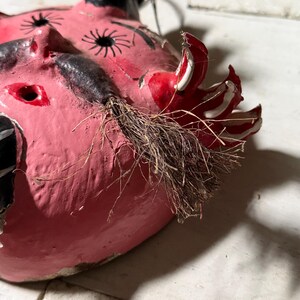 Puede incluir: Una m&aacute;scara de papel mach&eacute; rosa con detalles pintados en negro y blanco, un cuerno pintado en rojo y blanco y una barba de cerdas marrones.