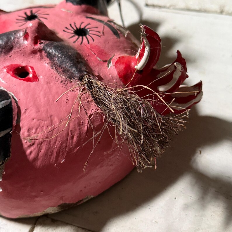 Puede incluir: Una m&aacute;scara de papel mach&eacute; rosa con detalles pintados en negro y blanco, un cuerno pintado en rojo y blanco y una barba de cerdas marrones.