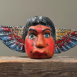 May include: A hand-carved wooden mask with a human face and wings. The face is painted red with blue eyes, black hair, and red lips. The wings are painted with blue, red, and yellow accents. The mask is displayed on a wooden surface.