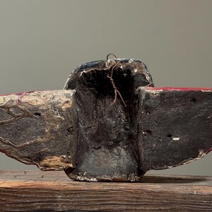 May include: A weathered, carved wooden mask with a dark, textured surface. The mask features wing-like extensions with red accents and a central cavity. A wire is attached at the top. The mask rests on a wooden surface.
