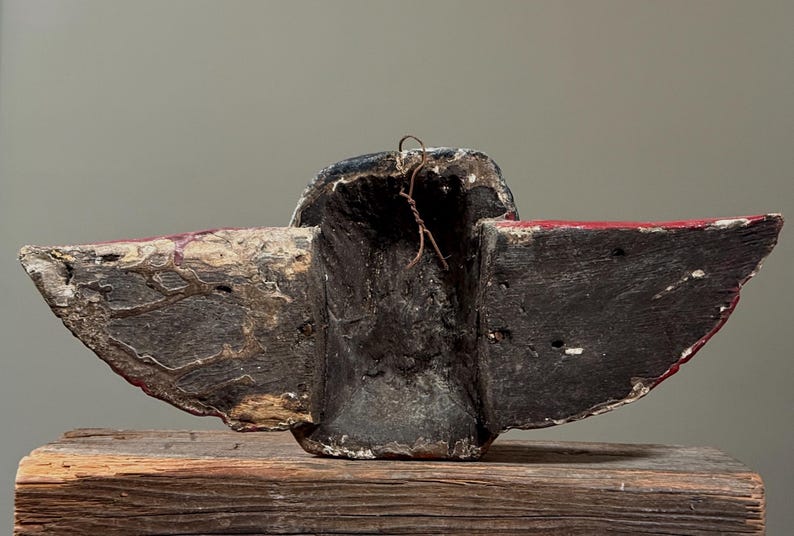 May include: A weathered, carved wooden mask with a dark, textured surface. The mask features wing-like extensions with red accents and a central cavity. A wire is attached at the top. The mask rests on a wooden surface.