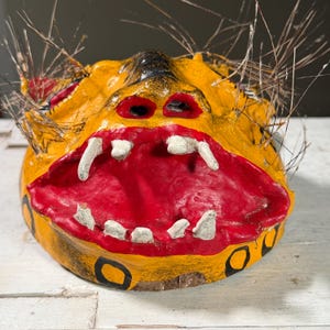 Puede incluir: Una m&aacute;scara de papel mach&eacute; amarilla y roja con una expresi&oacute;n de gru&ntilde;ido. La m&aacute;scara tiene detalles pintados de negro y dientes blancos. La m&aacute;scara est&aacute; decorada con rayas negras y amarillas y tiene una franja de plumas marrones alrededor de los bordes.