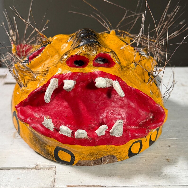 Puede incluir: Una m&aacute;scara de papel mach&eacute; amarilla y roja con una expresi&oacute;n de gru&ntilde;ido. La m&aacute;scara tiene detalles pintados de negro y dientes blancos. La m&aacute;scara est&aacute; decorada con rayas negras y amarillas y tiene una franja de plumas marrones alrededor de los bordes.