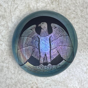 Può includere: Un fermacarte rotondo in vetro con un design nero e blu iridescente di un'aquila con le ali spiegate. L'aquila è delineata in nero e ha un design dettagliato di piume.
