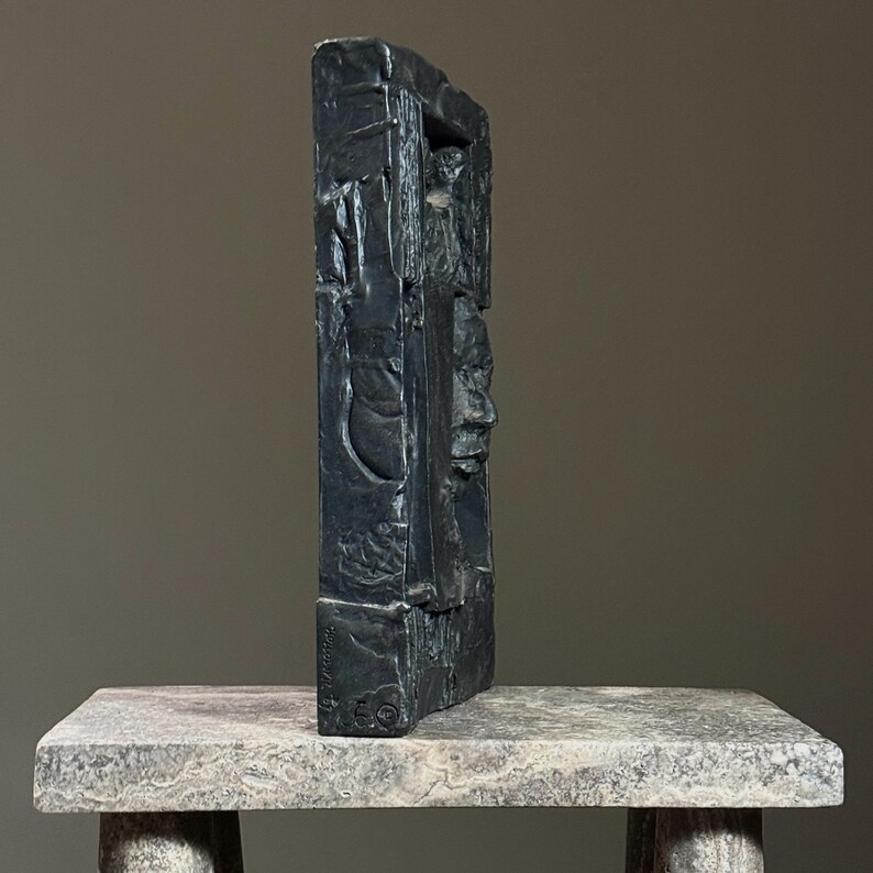 May include: Black abstract sculpture of a face with a textured surface. The sculpture is made of a dark material and has a rectangular shape. The sculpture is sitting on a light gray surface.