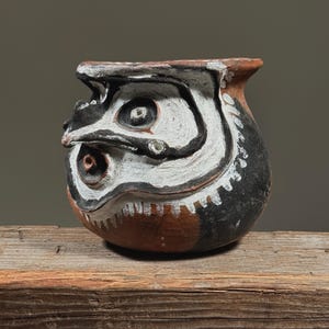May include: A small, round, hand-painted ceramic vessel with a stylized face design. The vessel features a black base, with white and brown accents. The face has large, round eyes and a beak-like nose. The vessel is sitting on a wooden surface.