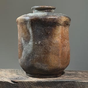 May include: A handcrafted, rustic ceramic vase with a cylindrical body and a slightly flared rim. The vase features a blend of brown, tan, and gray tones, with a textured surface. The vessel is displayed on a weathered wooden surface.
