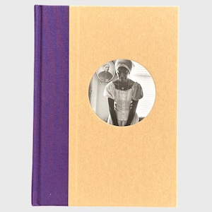 Puede incluir: Un libro de tapa dura con lomo morado y cubierta beige. Un recorte circular en la portada revela una fotografía en blanco y negro de una persona con pañuelo y vestido. Un retrato enmarcado es visible.