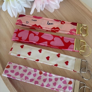 Op de afbeelding: Vier stoffen sleutelhangers met gouden of zilveren hardware. De sleutelhangers hebben verschillende patronen, waaronder harten, lippen en het woord "love". De sleutelhangers zijn in tinten roze, rood en wit.