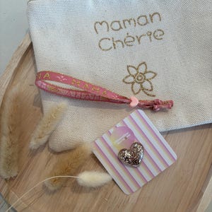 Peut inclure: Une pochette en toile crème brodée des mots "Maman Chérie" en or, un bracelet rose et une épingle en forme de cœur pailleté sur une carte à rayures, le tout disposé sur un plateau en bois. Des fleurs séchées complètent l'ensemble.