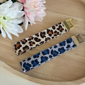 Op de afbeelding: Twee sleutelhangers met goudkleurige hardware. Eén heeft een bruin en beige luipaardprint, en de andere een blauwe en grijze luipaardprint. De sleutelhangers liggen op een houten dienblad met roze en witte bloemen.