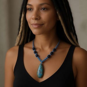 May include: A blue macrame necklace with a teardrop-shaped pendant. The pendant is a mix of blue and green hues. The necklace also features black beads and is worn against a black tank top.