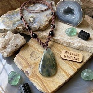 May include: A moss agate and macrame necklace with a teardrop-shaped pendant. The pendant is green and white. The necklace has black beads and a brown cord. The image also includes crystals and a wooden sign that says "MOSS AGATE & MACRAME."