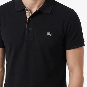 May include: Black polo shirt with short sleeves and a classic collar. The collar lining features a plaid pattern in beige, red, and black. A silver emblem is on the left chest.