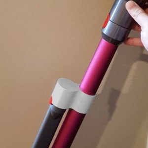Puede incluir: Una aspiradora inalámbrica con un diseño magenta y gris. La aspiradora tiene una varilla larga con un cabezal de limpieza desmontable. La empuñadura la sostiene una persona. La aspiradora está diseñada para la limpieza del hogar.