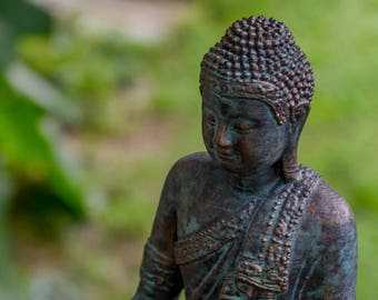 May include: A bronze-colored statue of a meditating Buddha. The statue is sitting in a meditative pose with hands clasped in the lap. The statue has a detailed face and intricate clothing.