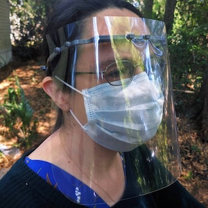 May include: A person wearing a clear plastic face shield with a blue surgical mask underneath. The face shield has a black headband and is secured with a strap around the back of the head.