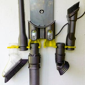 May include: A wall-mounted vacuum cleaner accessory organizer with a gray and yellow design. The organizer has slots for storing various attachments, including a hose, a crevice tool, and a dusting brush.