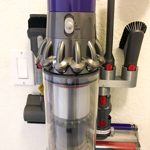 May include: A purple and silver cordless vacuum cleaner with a clear dustbin and a red button labeled "MAX". The vacuum is mounted on a wall-mounted storage rack with other attachments.