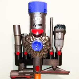 May include: A black wall mount with six slots for a Dyson vacuum cleaner and its attachments. The vacuum cleaner is blue and gray with a red button. The attachments include a crevice tool, a dusting brush, a combination tool, and a soft dusting brush.