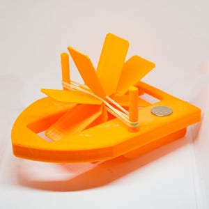 Puede incluir: Un barco de juguete de color naranja brillante impreso en 3D con una hélice propulsada por una banda elástica. El barco tiene un pequeño imán en la parte inferior.
