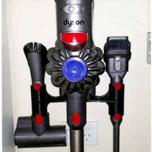 May include: A wall-mounted storage rack for a Dyson vacuum cleaner and its attachments. The rack is black and has two red dots on each side. The vacuum cleaner is gray and red, and the attachments are gray. The rack is mounted on a white wall.