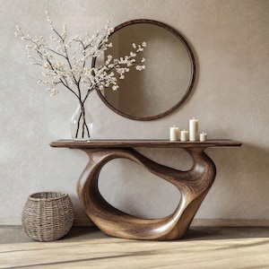 Puede incluir: Una consola de madera moderna y escultural con un diseño abierto y fluido. Un espejo redondo cuelga sobre la mesa, y un jarrón de cristal con flores blancas está encima. Varias velas blancas y una cesta de mimbre completan la decoración.