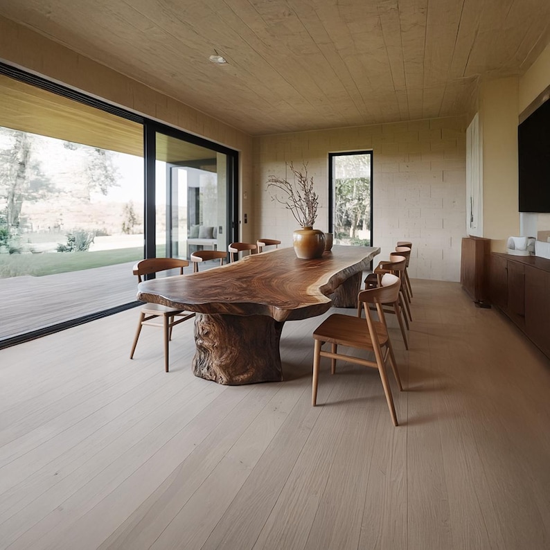 May include: A large, live-edge wooden dining table with a natural, organic shape. Surrounding the table are several wooden chairs. The room has light-colored wood flooring and a large window overlooking a natural landscape.