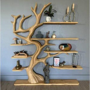 K&ouml;nnte beinhalten: Ein einzigartiges, baumf&ouml;rmiges Holzregal mit mehreren unterschiedlich gro&szlig;en Regalb&ouml;den. Die Regale beherbergen dekorative Gegenst&auml;nde wie Vasen, Figuren und eine kleine Topfpflanze. Das Regal ist hellbraun lackiert und an einer grauen Wand befestigt.