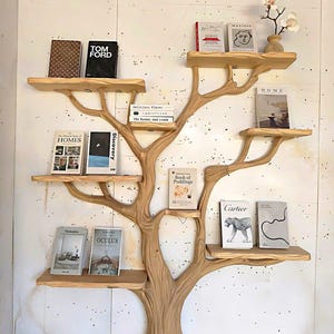 May include: A wooden tree-shaped bookshelf with multiple shelves holding various books. The shelves are light brown, and the tree trunk is a similar color. Books include titles like "TOM FORD," "HOMES," and "OCULUS." A small vase with flowers sits on the top shelf.