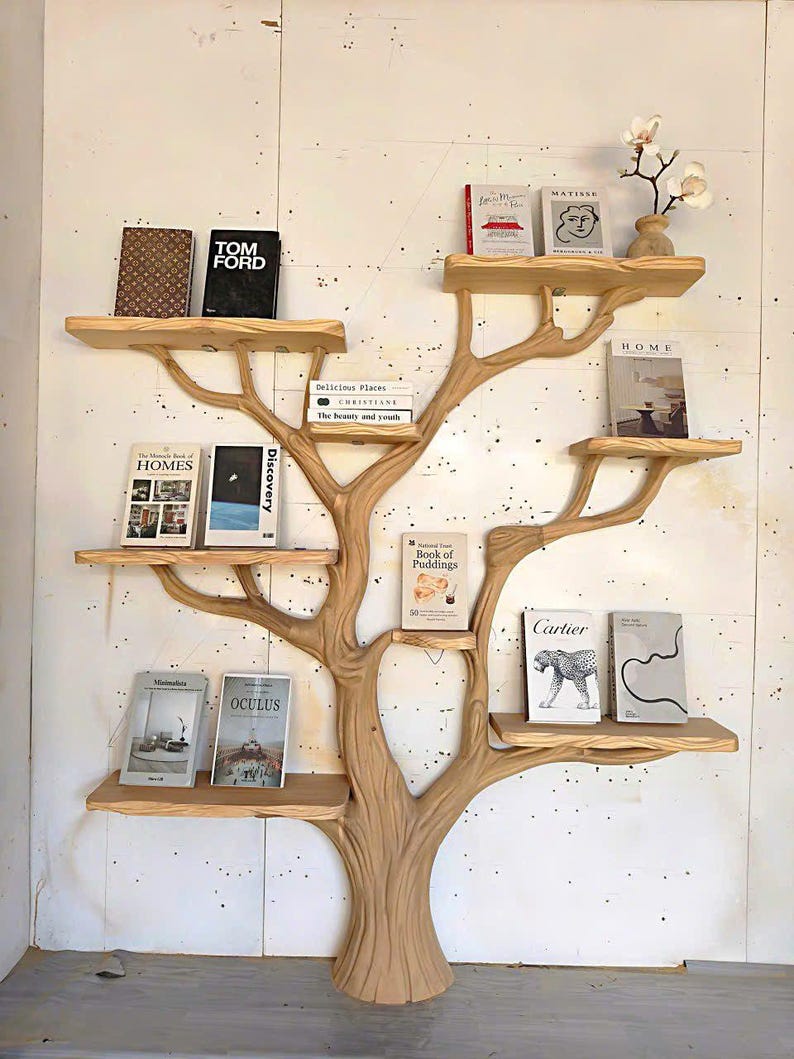 May include: A wooden tree-shaped bookshelf with multiple shelves holding various books. The shelves are light brown, and the tree trunk is a similar color. Books include titles like "TOM FORD," "HOMES," and "OCULUS." A small vase with flowers sits on the top shelf.